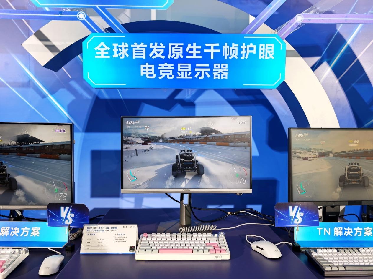 Monitor de computador com tela ligada exibindo jogo de corrida na neve com buggy off-road, posicionado sobre bancada azul entre dois outros monitores. Abaixo do monitor há um teclado mecânico branco com teclas rosa e cinza da marca AOC e um mouse branco. Placas azuis com a inscrição VS e uma placa informativa branca aparecem na frente, sob letreiro azul suspenso com texto em chinês