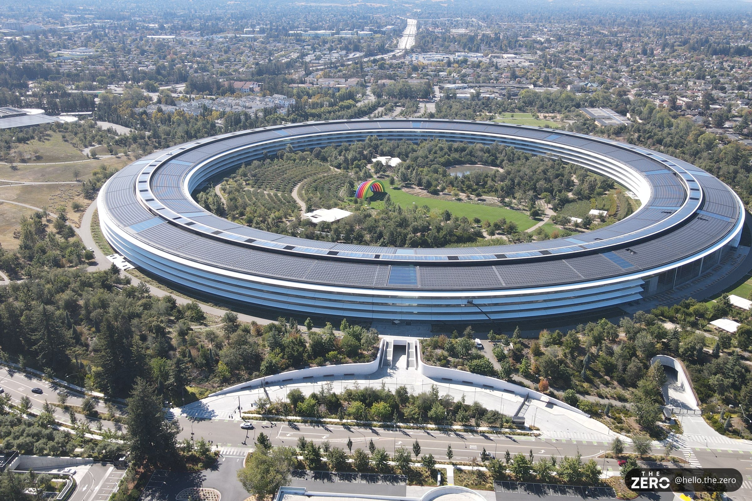 Vista aérea do Apple Park, em Cupertino, com o anel circular de vidro no centro do terreno, árvores ao redor e uma estrada no canto inferior esquerdo