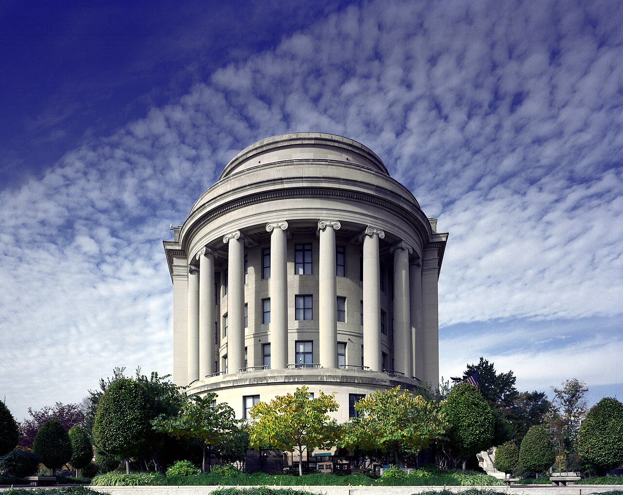 Fachada semicircular do Edifício da Federal Trade Commission em Washington, com colunas jônicas clássicas de pedra clara no topo, janelas escuras dispostas em vários andares, copa de árvores com folhagem verde e amarelada na base e céu azul coberto por nuvens brancas onduladas