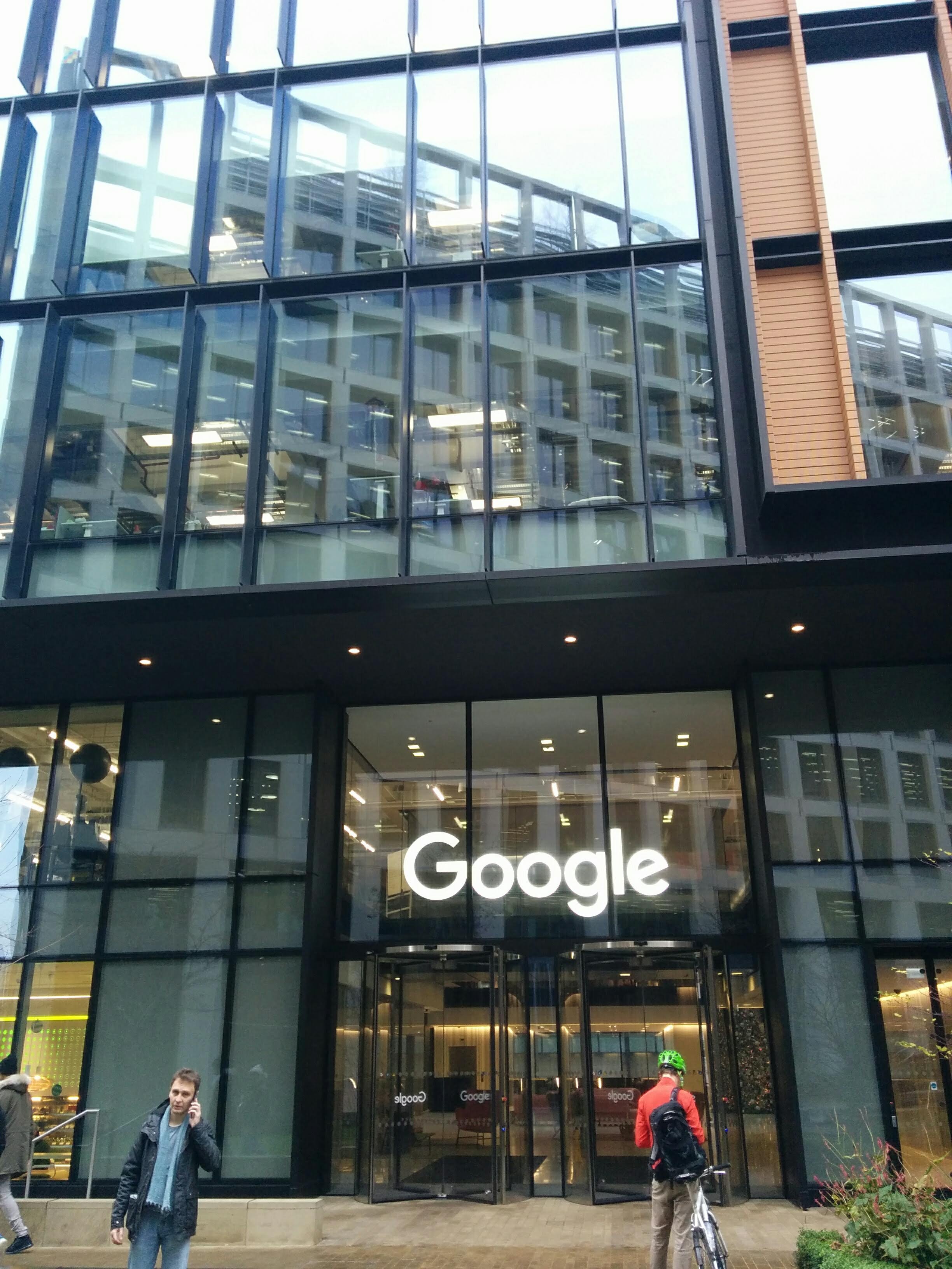 Entrada envidraçada de edifício comercial moderno com letreiro luminoso branco Google sobre portas giratórias de vidro. À esquerda, um pedestre de casaco escuro, cachecol azul claro e calça jeans fala ao celular. À direita, um ciclista de costas com jaqueta vermelha, capacete verde-limão e mochila preta segura uma bicicleta prateada. Acima da entrada, a fachada envidraçada reflete um prédio com janelas quadradas e traz acabamento lateral em painéis verticais de madeira clara