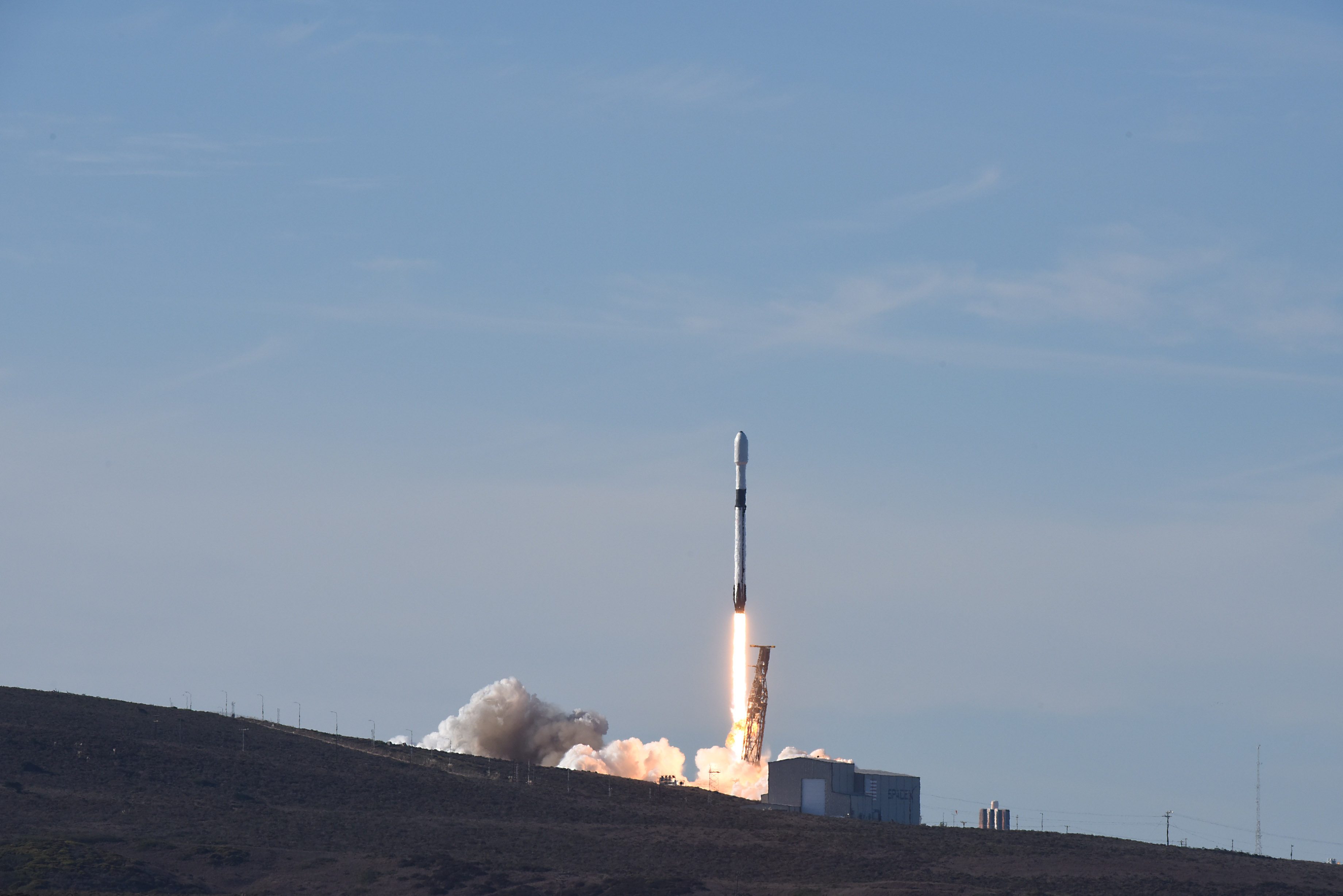Foguete Falcon 9 branco e preto em ascensão vertical após o lançamento, expelindo labaredas alaranjadas e fumaça branca densa sobre a base costeira, com colina escura em primeiro plano e céu azul ao fundo