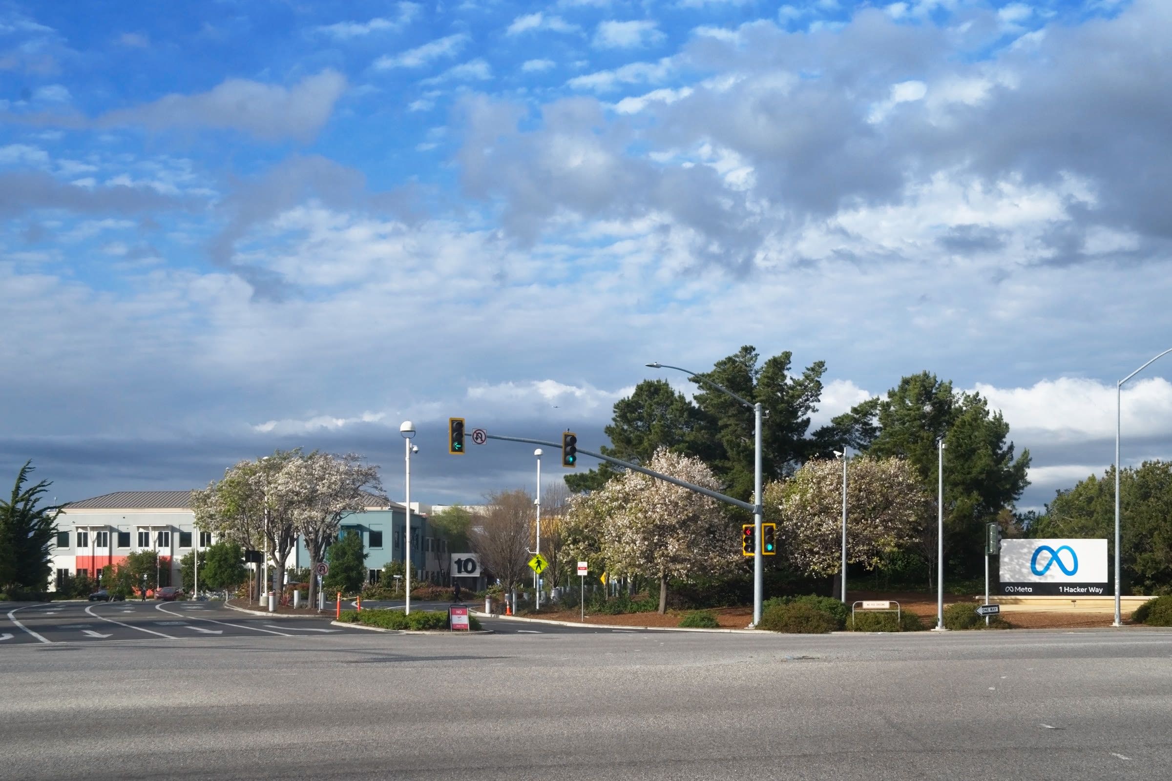 Letreiro branco tridimensional com as letras META sobre a grama, em frente a um prédio de vidro que mostra o símbolo do infinito e a palavra META, com uma avenida de várias faixas, carros, semáforos, árvores e céu azul com nuvens