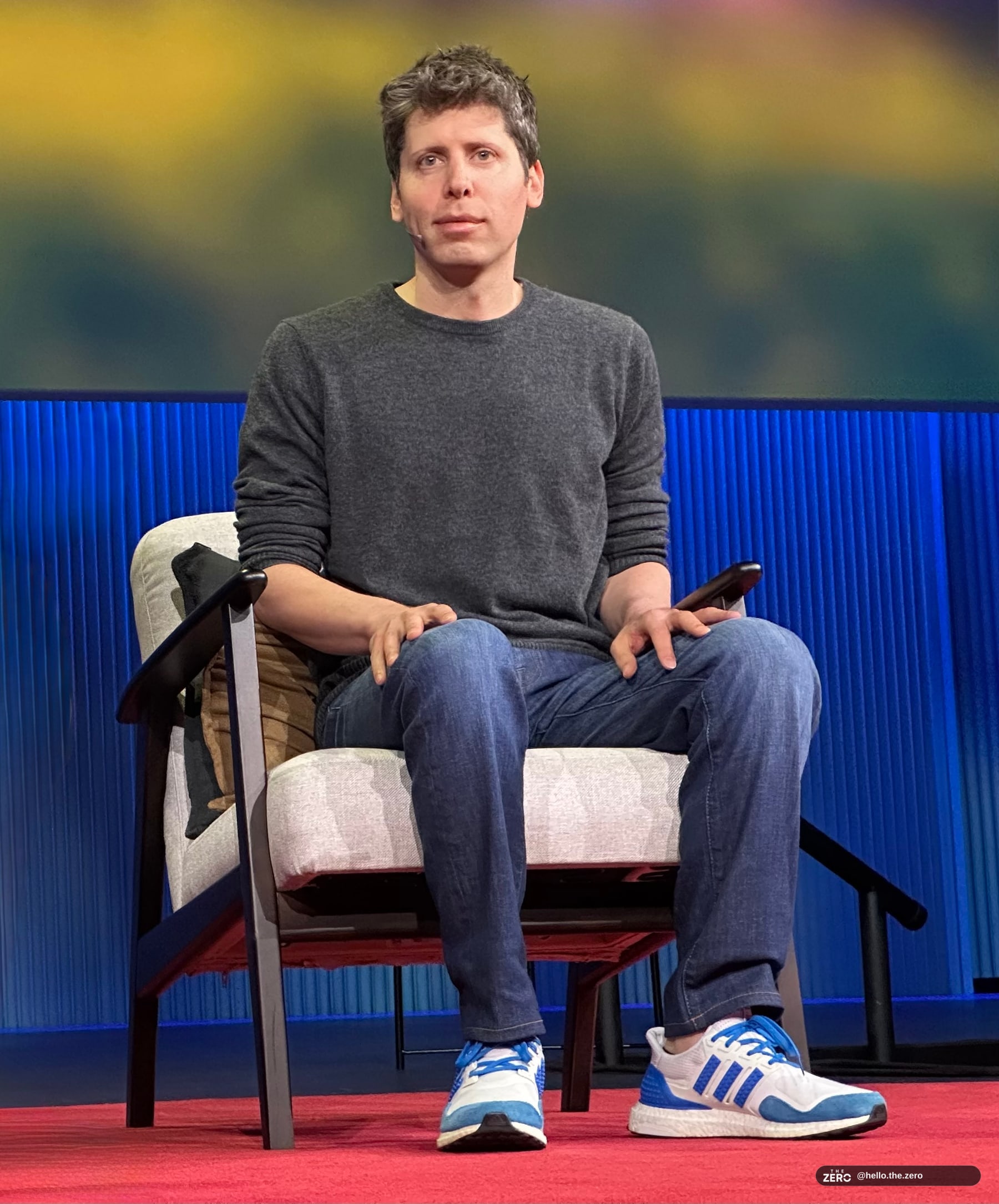 Sam Altman no palco do TED, de jaqueta escura e camisa clara, gesticula com a mão direita; a plateia aparece em silhueta e as letras TED estão no fundo
