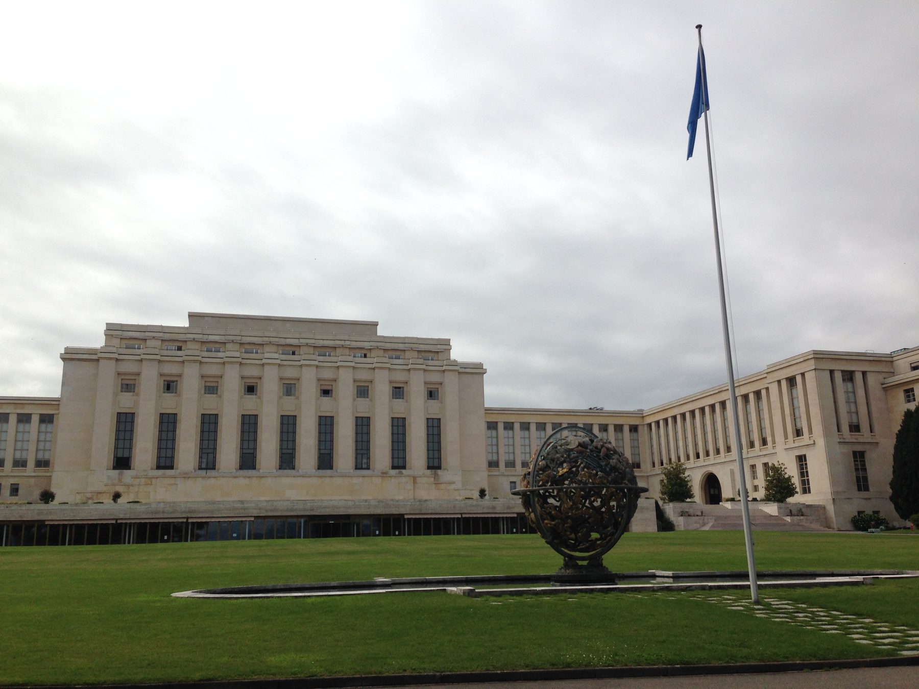 Fachada clara do Palais des Nations vista do lado do lago, com bandeiras, árvores e o céu azul