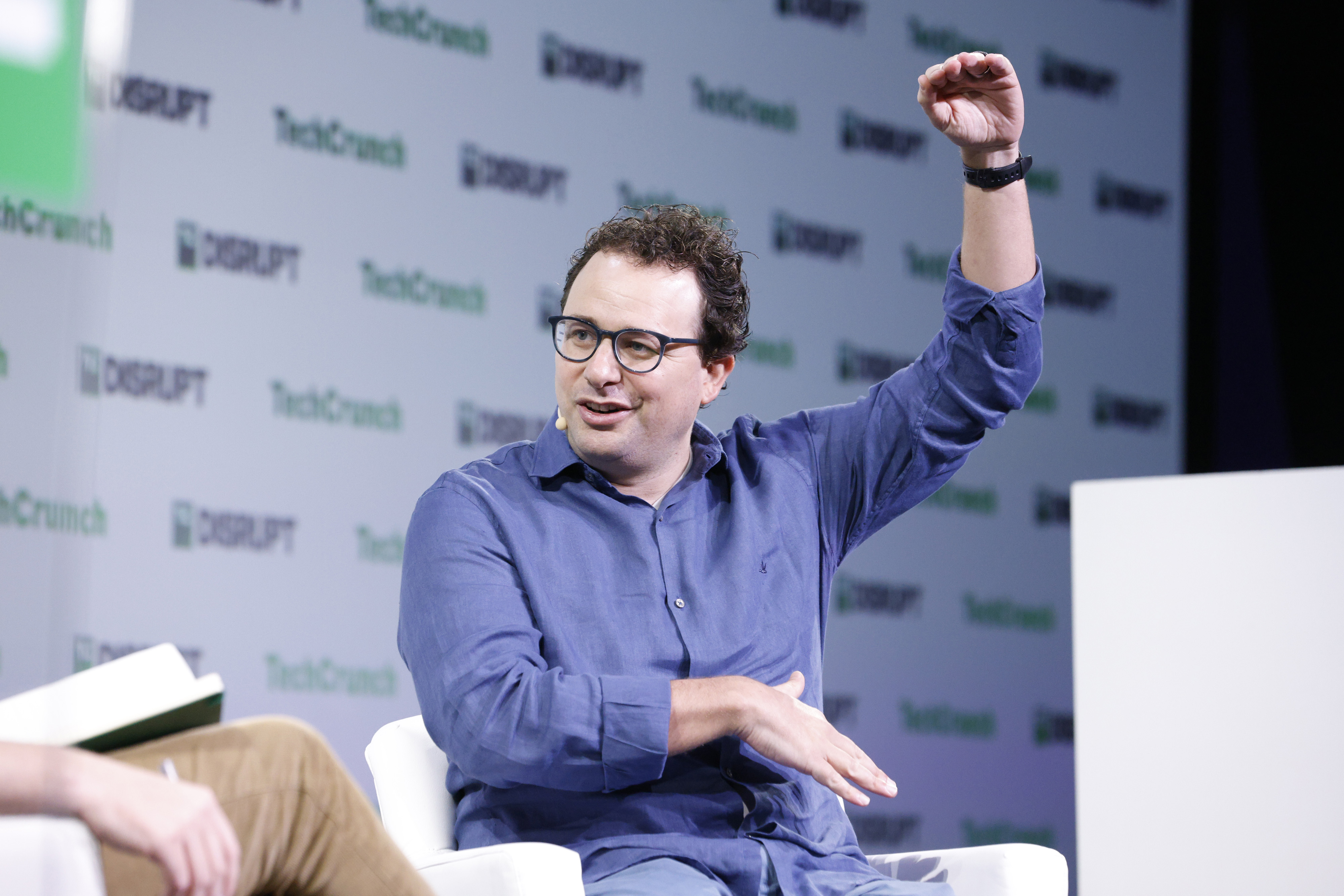 Dario Amodei, de cabelo curto, barba rala e suéter cinza escuro, em pé num palco escuro, com microfone preto na mão direita e a mão esquerda aberta, diante de uma tela em que se lê TechCrunch e Disrupt